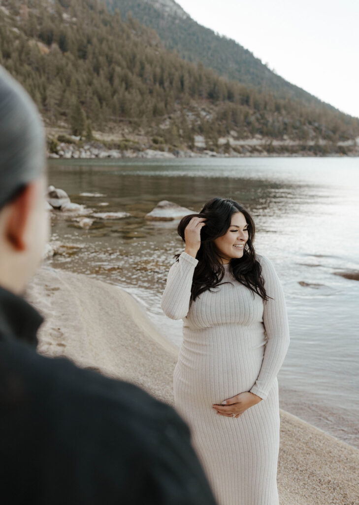 Pregnant woman holding hand over stomach while smiling and playing with hair with husband watching on Lake Tahoe beach