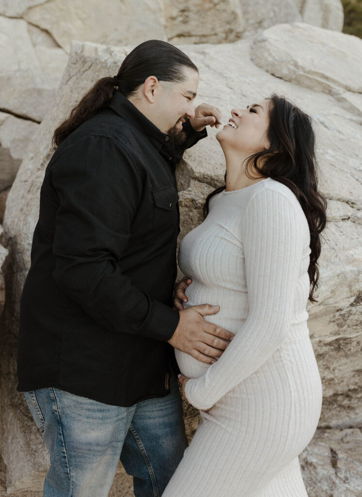 Couple smiling at each other while leaning on large rocks and keeping hands on pregnant woman's belly in Lake Tahoe