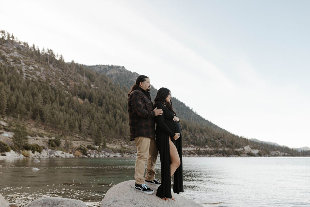 Couple standing on large rock while looking out over Lake Tahoe while pregnant woman keeps hand on belly 