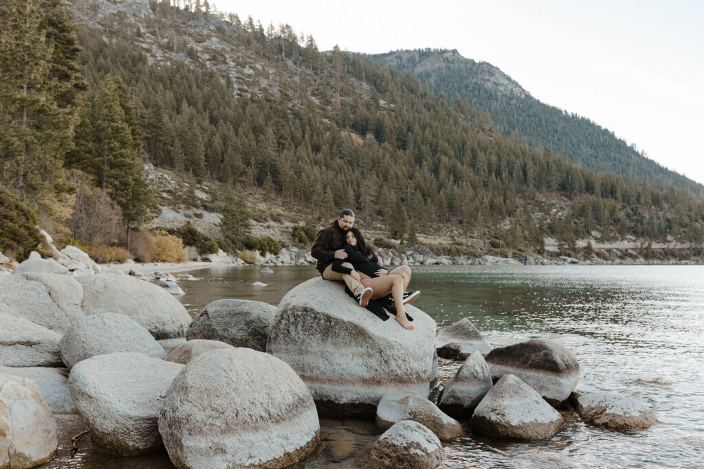 Couple lying on large rock together while surrounded by water at Lake Tahoe with mountains and trees in background