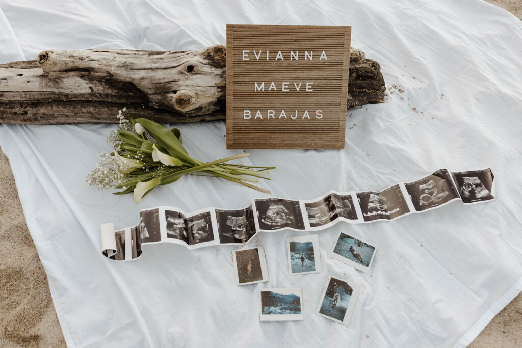 Baby announcement photos laid out on white blanket with white flowers and wooden sign on Lake Tahoe beach