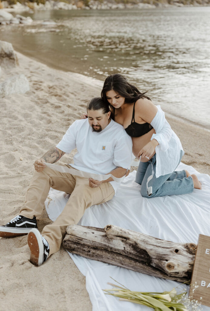 Couple looking over baby announcement photos together while sitting on white blanket together on sandy beach in Lake Tahoe