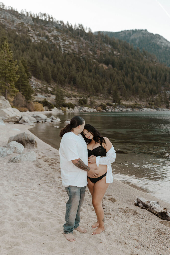 Couple standing together on sandy beach while husband kisses forehead and both keep hand on pregnant woman's belly in Lake Tahoe