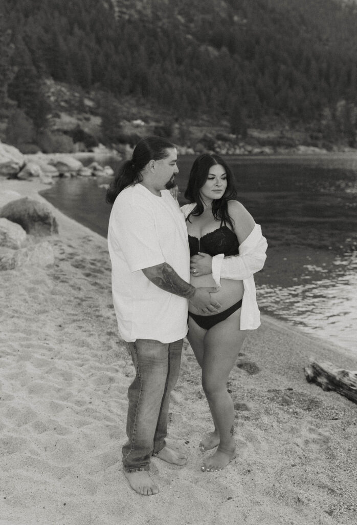 Couple standing together barefoot in the sand on beach in Lake Tahoe while both keep a hand on pregnant woman's belly