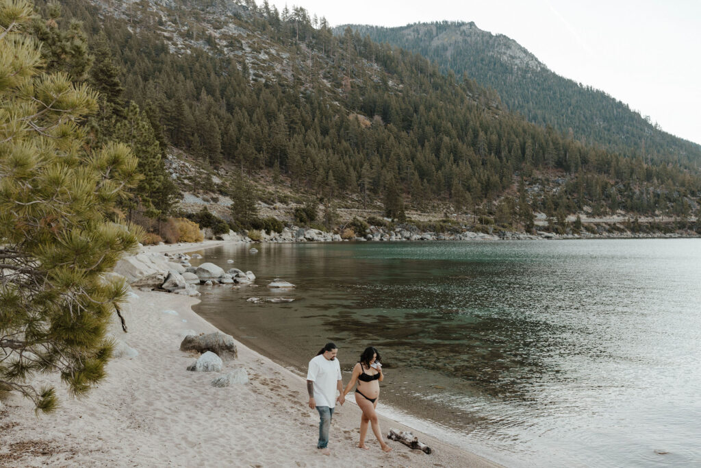 Pregnant woman holding hands with husband and walking down sandy beach together in Lake Tahoe with lake and trees in background