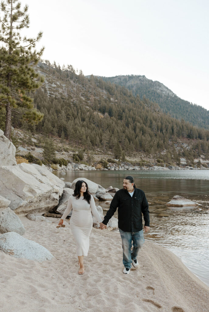 Couple holding hands while walking down sandy beach together in Lake Tahoe with lake and trees in background