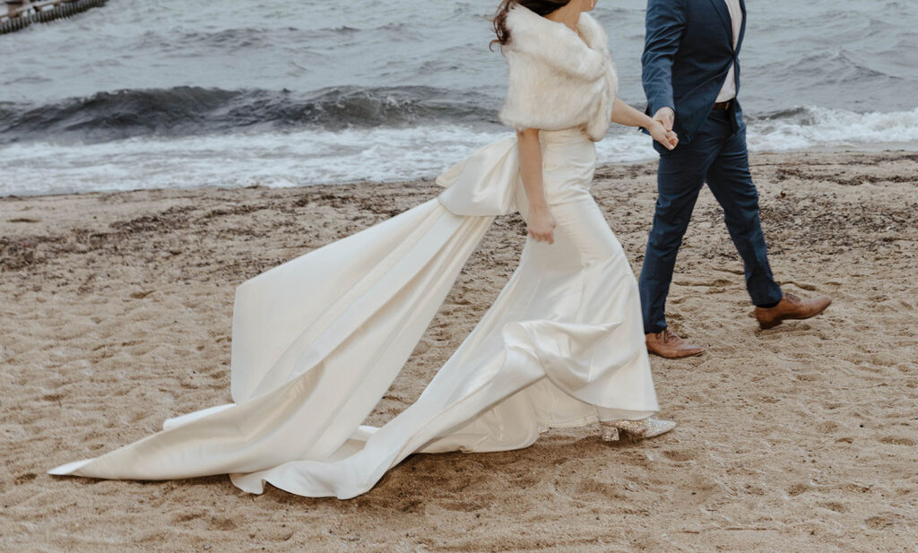 bride and groom walking on the beach as the wind blows her wedding dress 