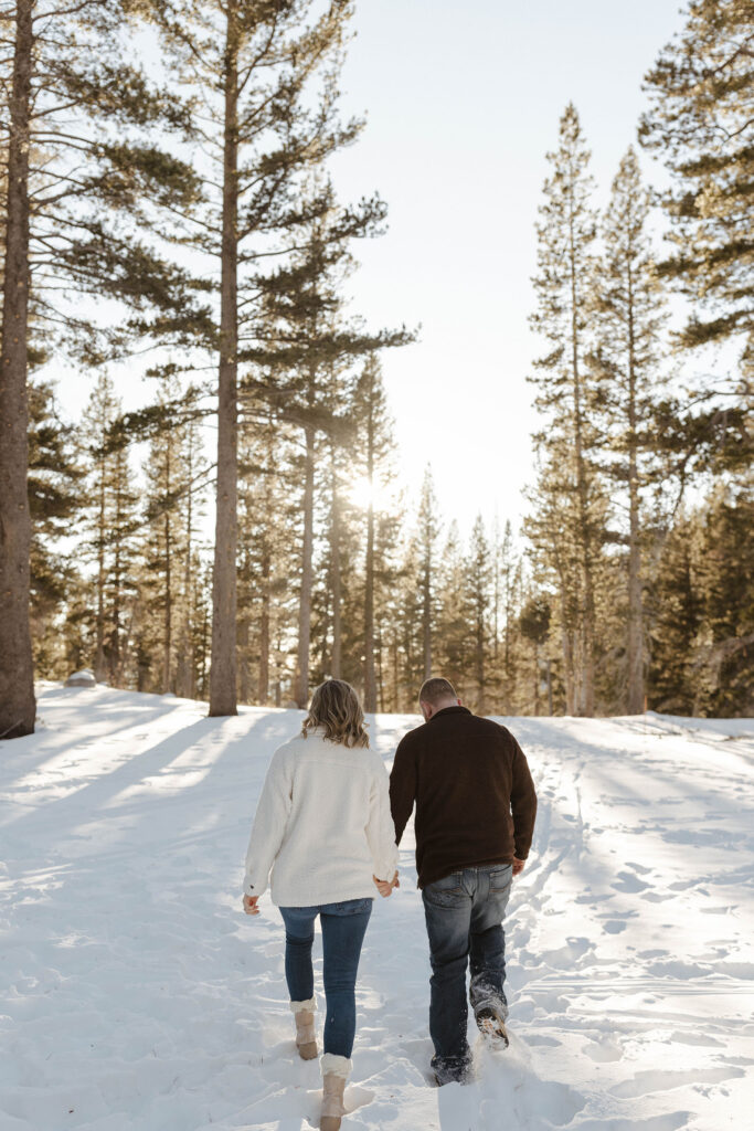 Couple holding hands while walking through snow together toward pine trees in Lake Tahoe