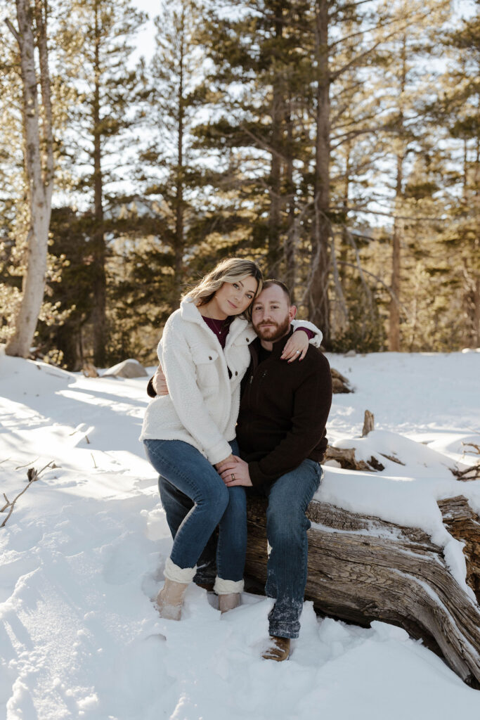 Couple sitting on log together while holding each other and looking at camera while surrounded by snow in Lake Tahoe