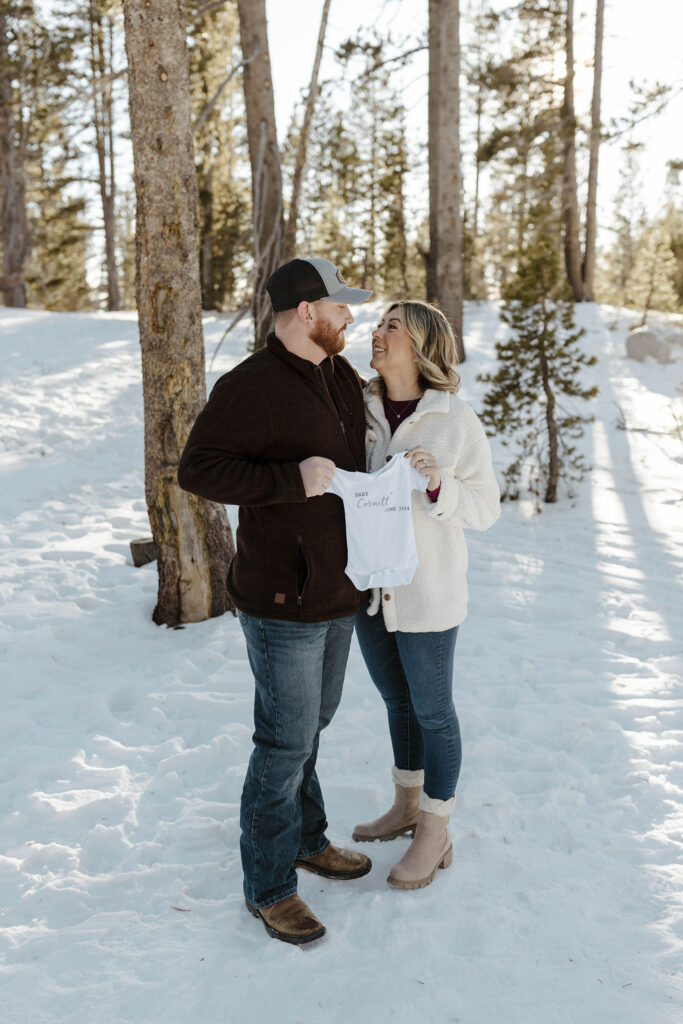 Couple holding up baby clothes while smiling at each other and standing in snow at Lake Tahoe