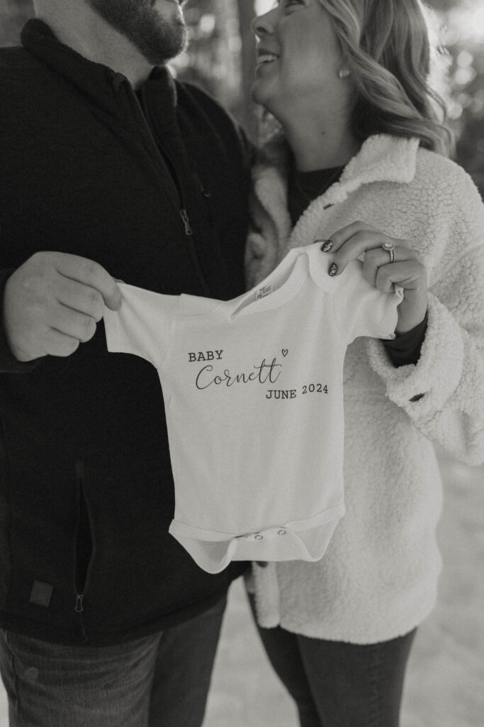 Close up of couple holding baby clothes announcing pregnancy while smiling at each other in Lake Tahoe