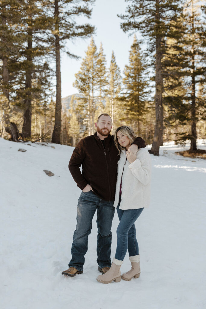 Couple holding each other while facing camera and standing in snow with pine trees in background at Lake Tahoe
