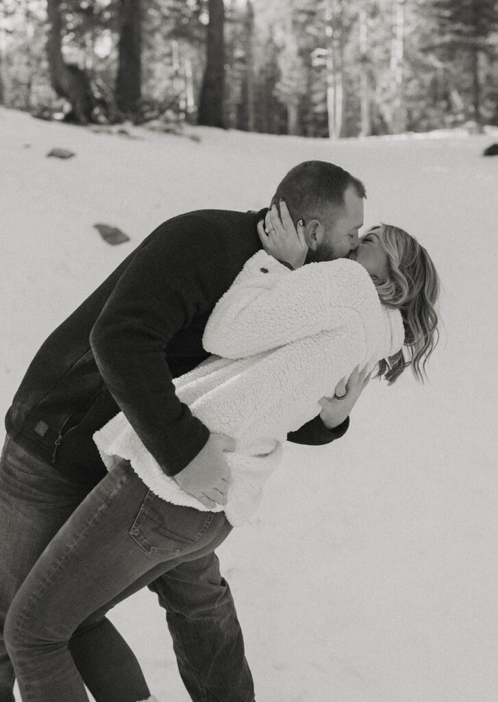 Couple holding each other while kissing and standing in the snow with pine trees in background