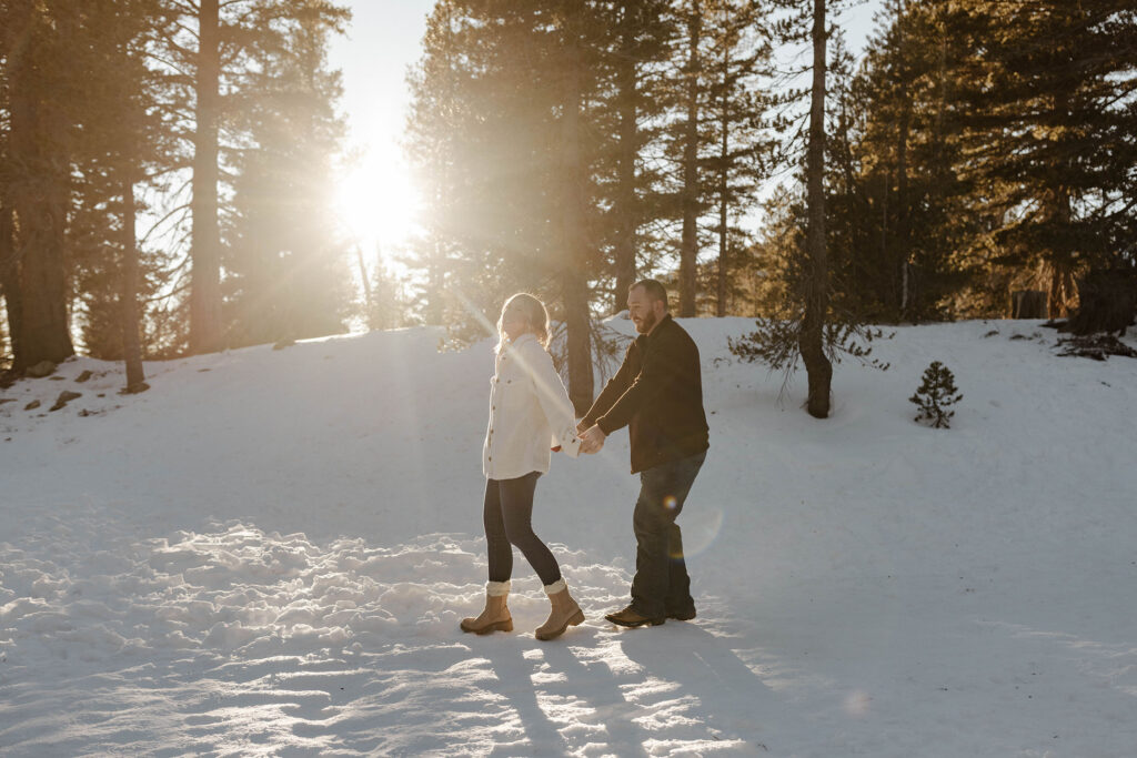 Couple holding hands while woman leads man through the snow with sun shining through the pine trees in Lake Tahoe