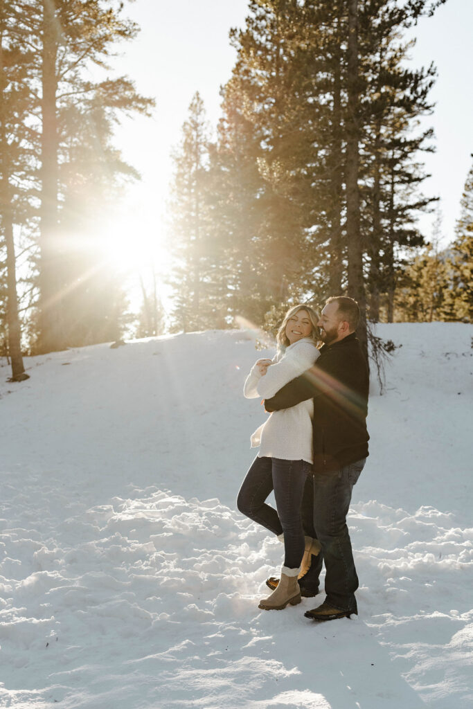 Couple hugging each other while smiling and standing in the snow with sun shining through pine trees in background