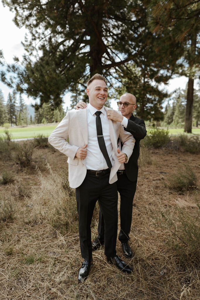Groom's dad helping groom put on wedding jacket while smiling and standing in front of pine trees in Old Greenwood