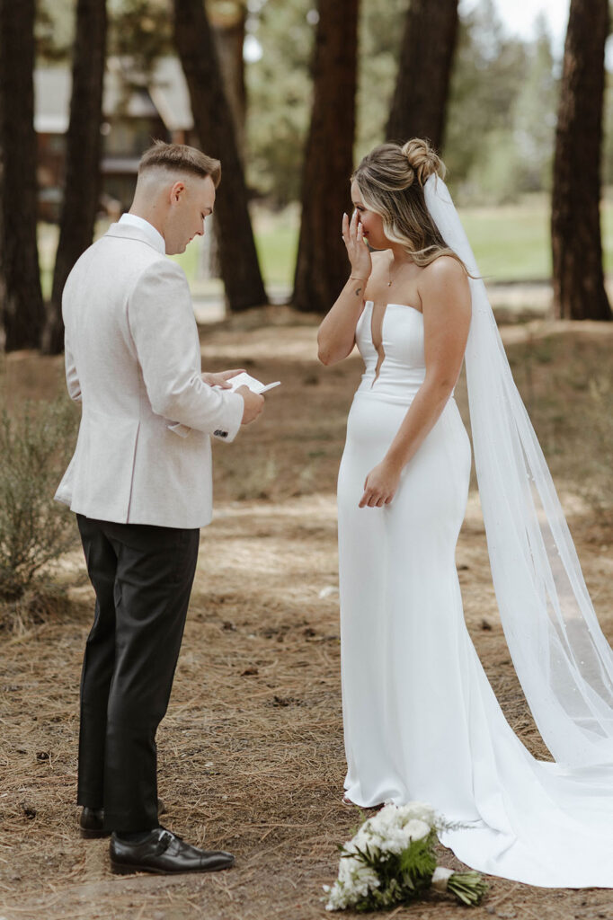 Bride emotional while groom reads wedding vows to her in front of pine trees at Timbers at Old Greenwood