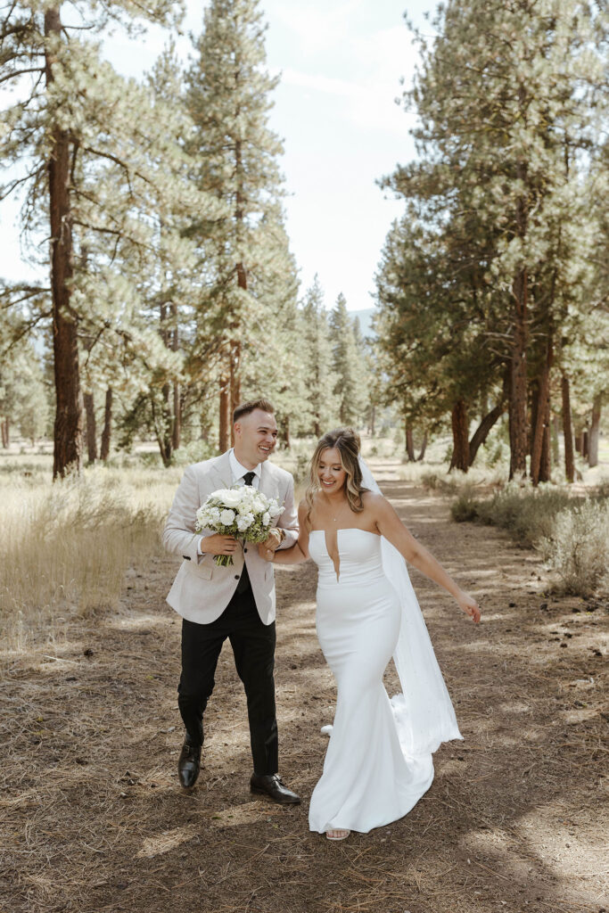 Wedding couple laughing while walking down dirt trail together with pine trees in background at Old Greenwood