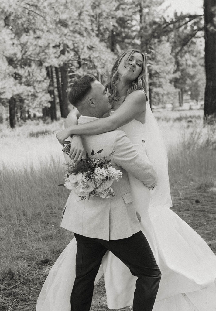 Groom picking wedding bride up while she holds floral bouquet with pine trees in background at Old Greenwood