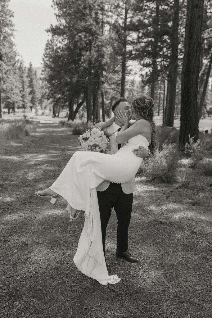 Wedding groom carrying bride while they kiss on a dirt trail with pine trees in background at Timbers at Old Greenwood