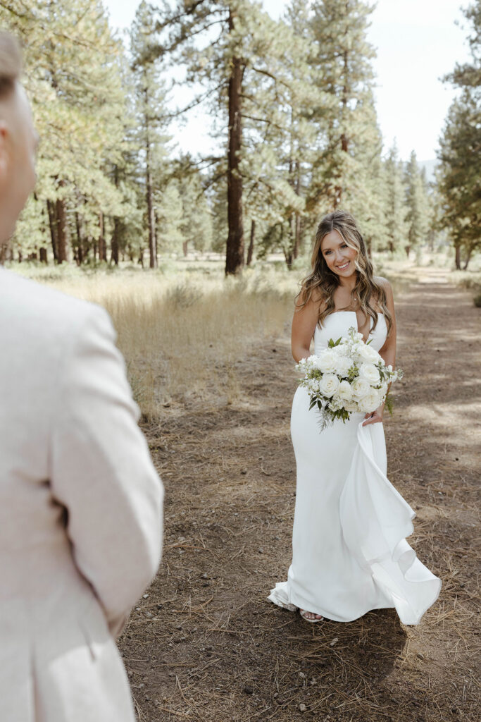 Wedding bride standing and holding floral bouquet while smiling at groom with pine trees in background at Old Greenwood