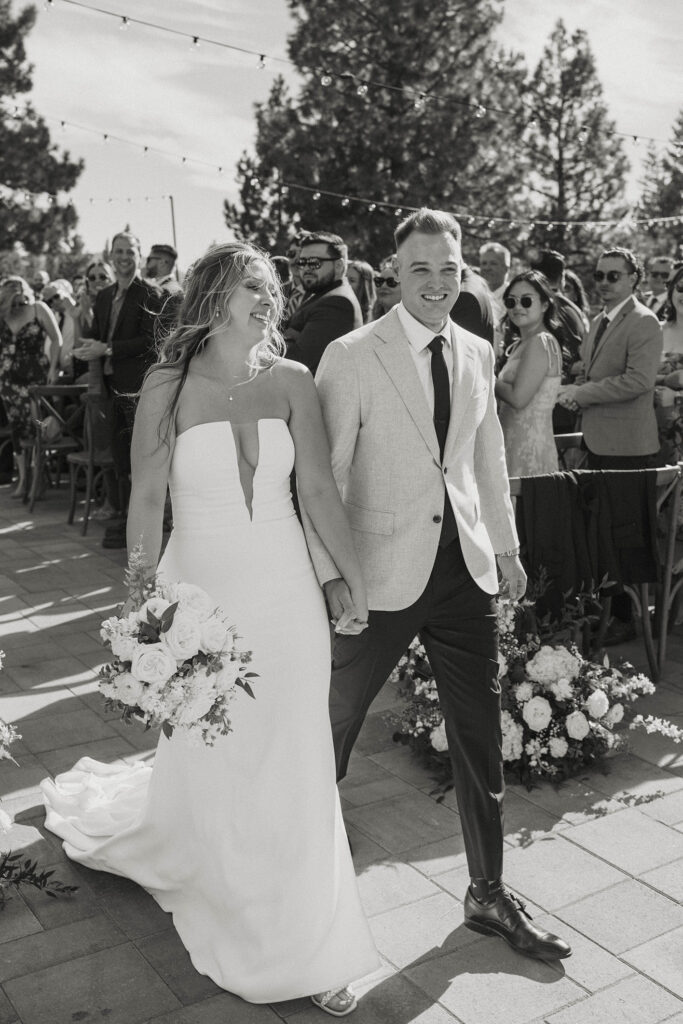 Wedding couple holding hands while smiling and walking down aisle out of ceremony while guests smile and clap at Timbers at Old Greenwood