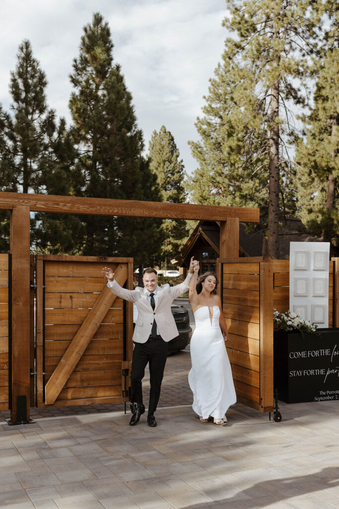 Wedding couple holding hands in air and celebrating during grand entrance while walking out of wooden gate into reception at Old Greenwood