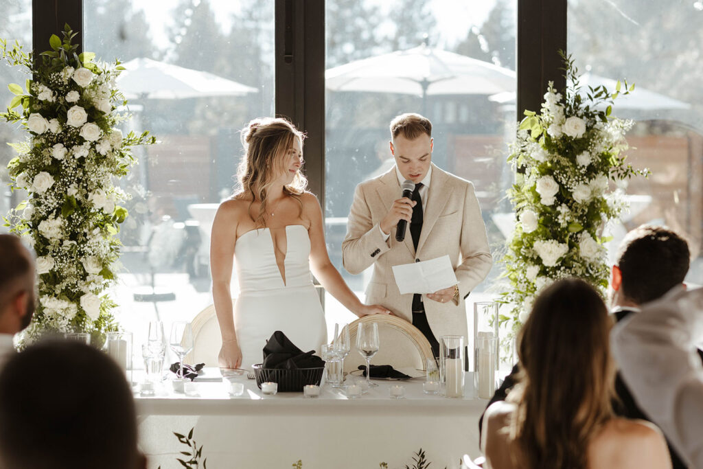 Groom reading speech while holding microphone and standing next to bride at head table during wedding reception at Timbers at Old Greenwood