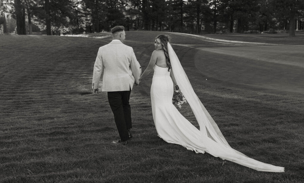Wedding couple walking across grassy field while holding hands and bride looks back at camera at Old Greenwood