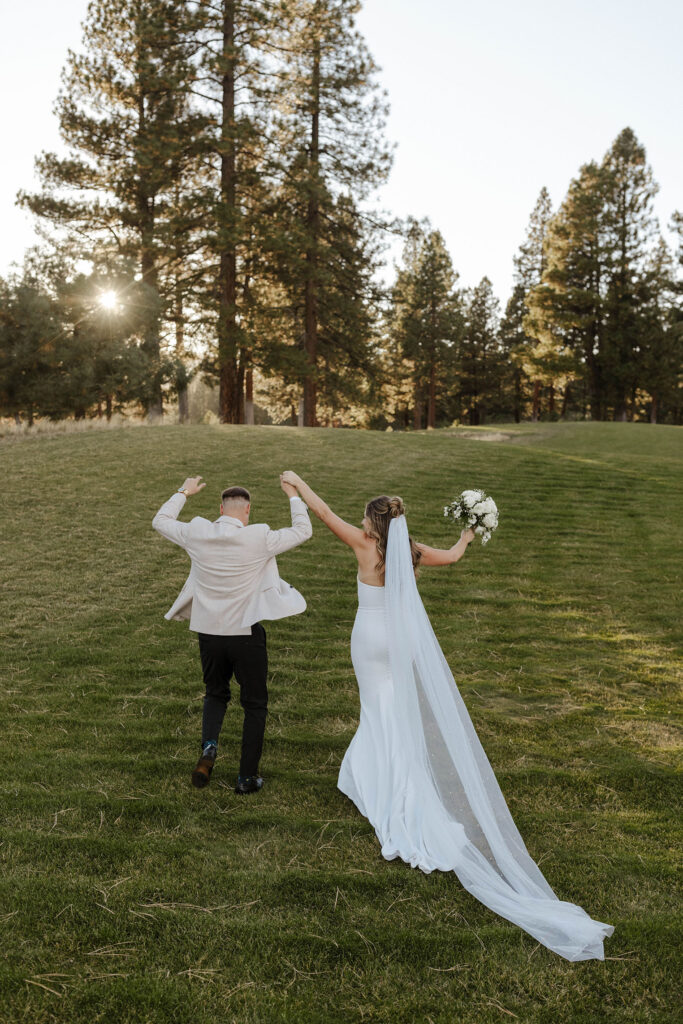Wedding couple celebrating while holding hands and walking away from camera across grassy field toward pine trees at Timbers at Old Greenwood