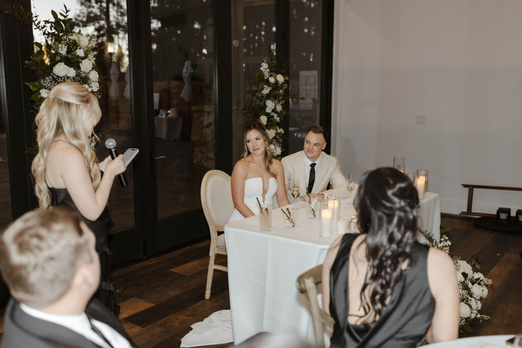 Bride and groom sitting at head table listening to speeches and smiling during wedding reception at Timbers at Old Greenwood
