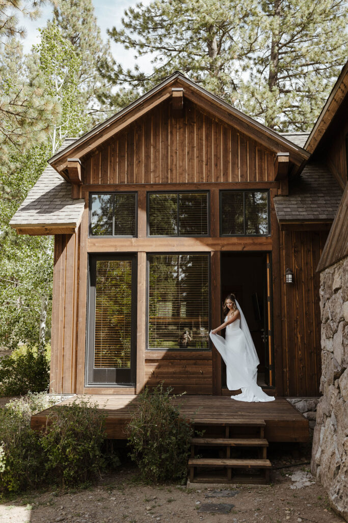 Bride in wedding dress holding dress as she walks in front of wooden building outside with large windows at Old Greenwood