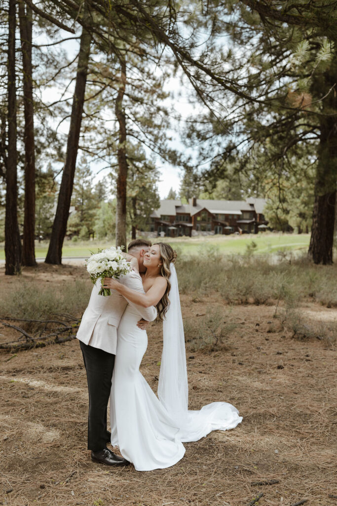 Wedding couple hugging while outside in front of pine trees with large wooden building in background at Old Greenwood