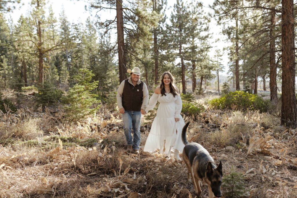 Engagement couple holding hands while walking in dry grass behind dog with pine trees in background in Truckee
