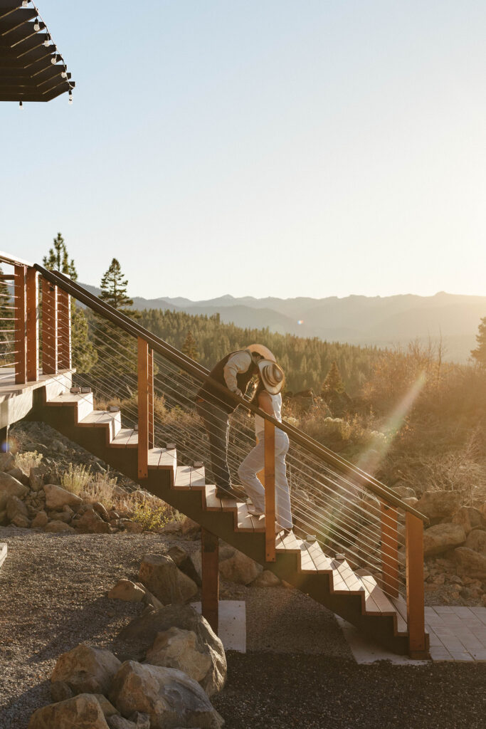 Engagement couple standing on staircase and kissing each other while wearing cowboy hats with lots of pine trees in background in Truckee