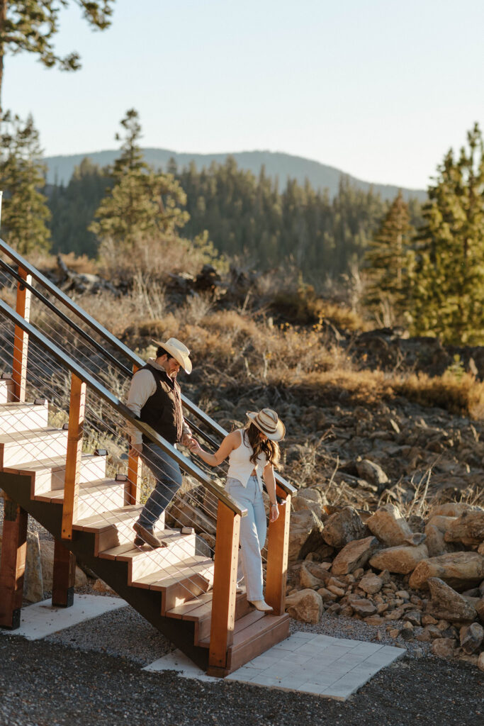 Woman holding fiancé's hand and leading him down staircase while both wear cowboy hats in Truckee