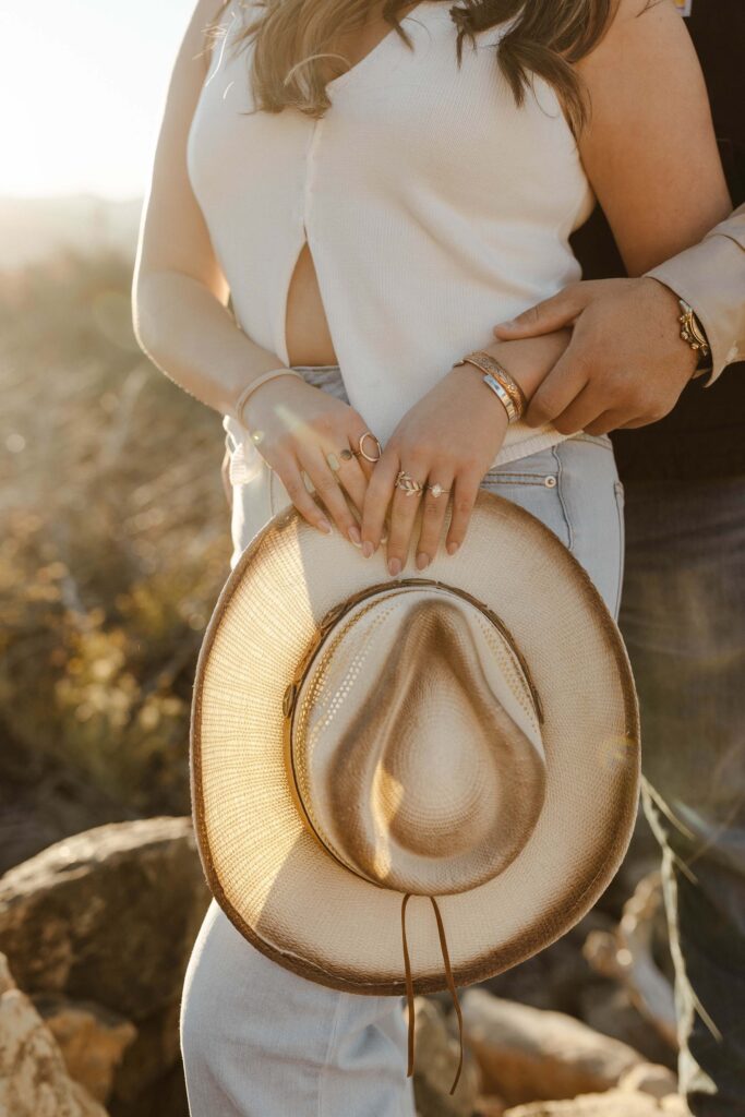 Close up of woman holding cowboy hat with engagement ring on finger and fiancé hugging her from behind in Truckee