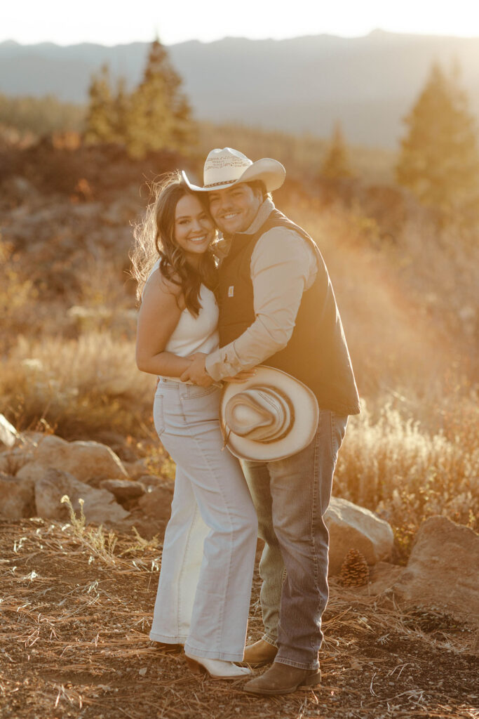 Engagement couple hugging each other while smiling at camera with rocks and dry grass in background in Truckee