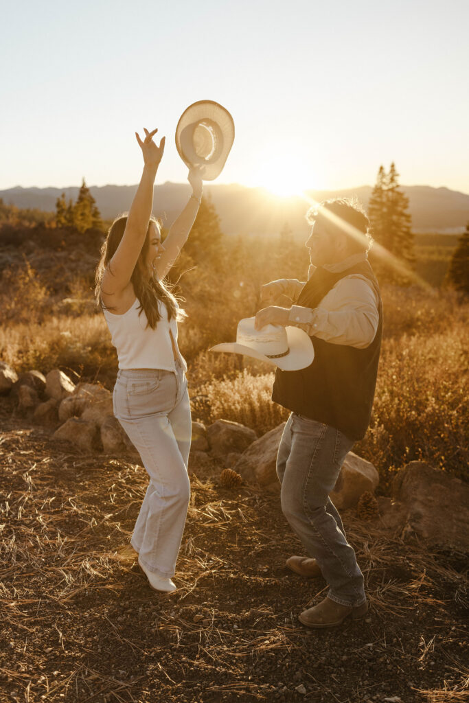 Engagement couple celebrating while holding cowboy hats with dry grass and rocks in background in Truckee