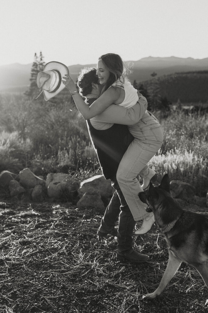 Man picking up fiancé and hugging her while she is holding cowboy hat with shrubs in background in Truckee