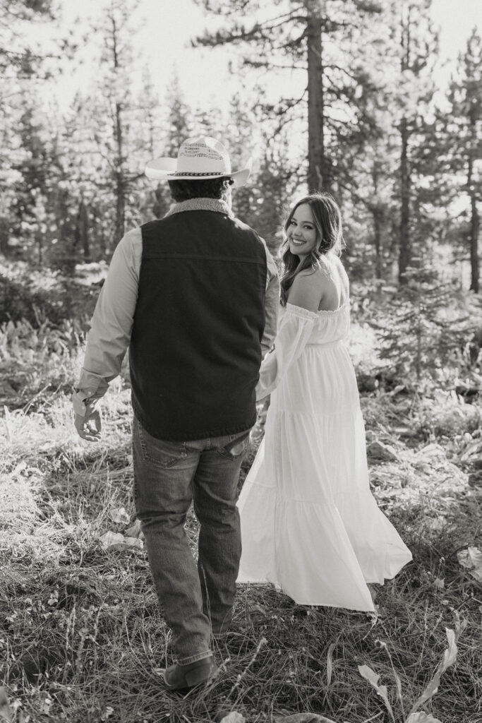 Woman looking over shoulder at camera as engagement couple hold hands and walk along dry grass in Truckee