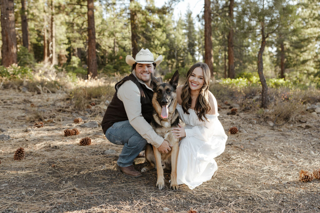 Engagement couple kneeling by their dog and holding him while all smile at the camera with pine trees in background in Truckee