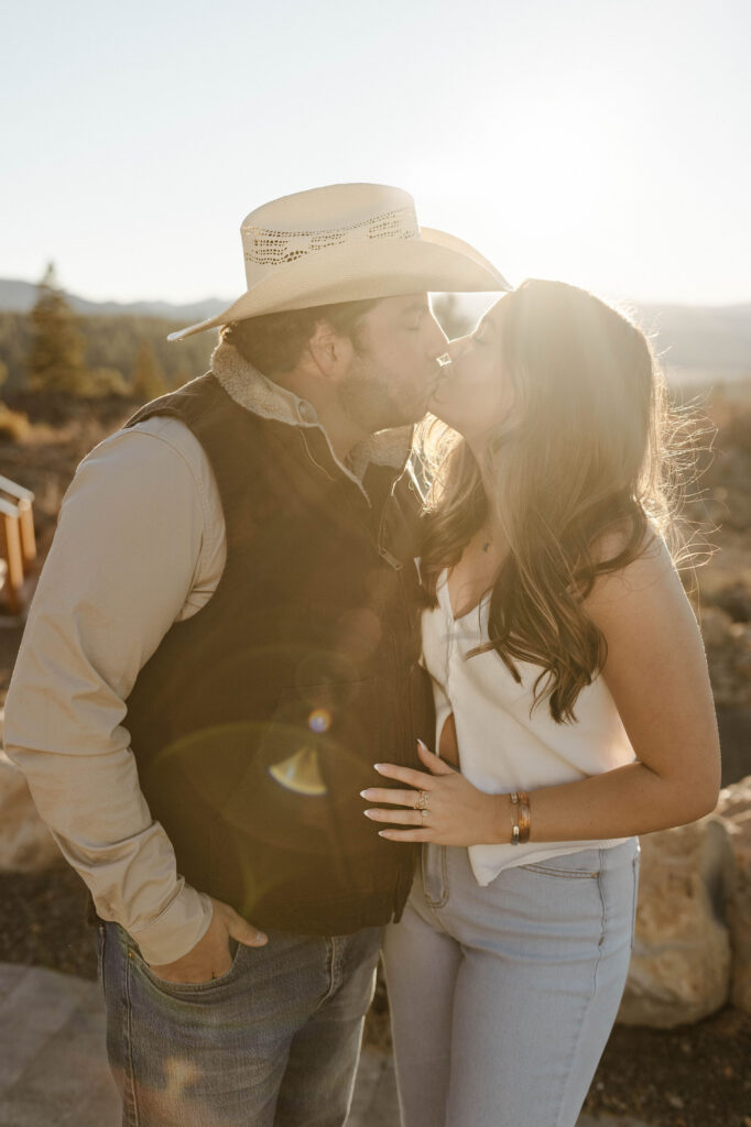 Engagement couple holding each other and kissing while sun glares in background in Truckee