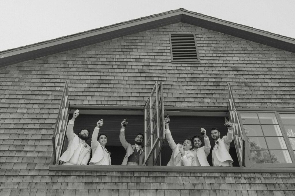 Wedding groom and groomsmen holding up beers while leaning out window above camera at Valhalla in Lake Tahoe