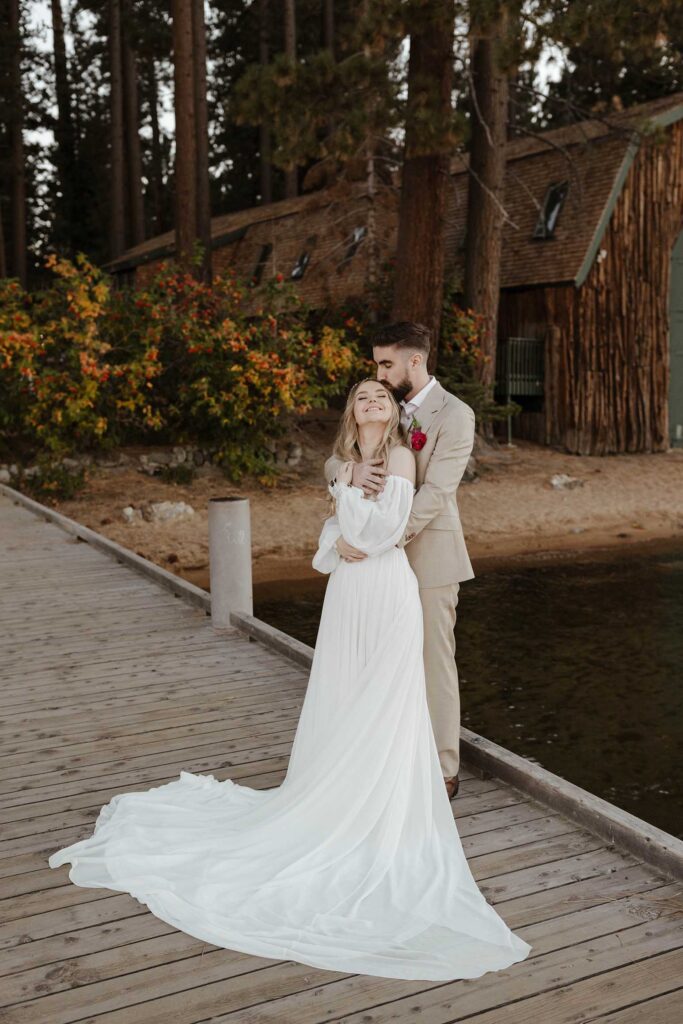 Groom hugging bride from behind while kissing forehead and standing on wooden dock at Valhalla in Lake Tahoe