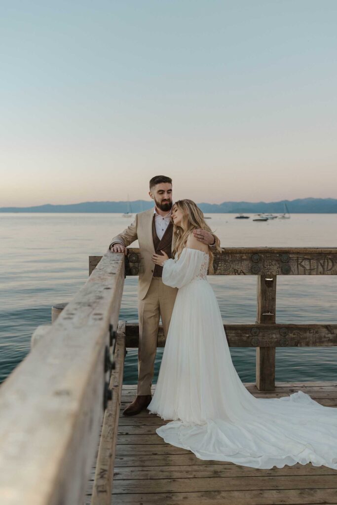 Wedding couple hugging each other while leaning on wooden dock railing on Lake Tahoe at Valhalla