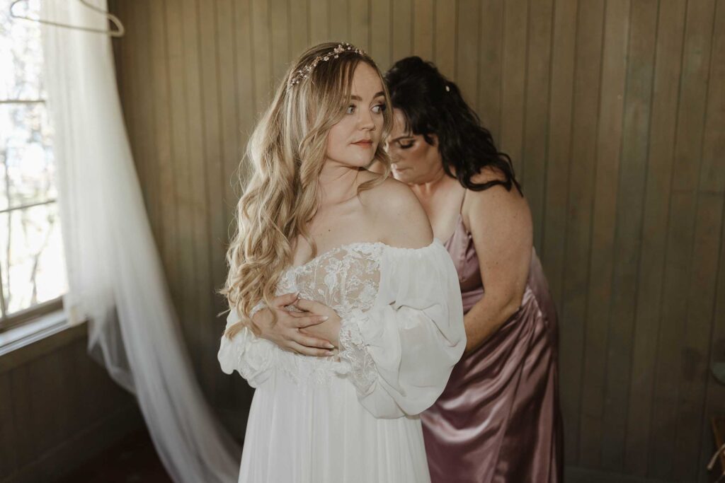 Bride's mom helping bride put on wedding dress while bride looks to side inside Valhalla