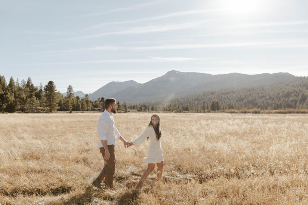 Couple walking through golden field while holding hands and woman looks back at man in Lake Tahoe with mountains in background