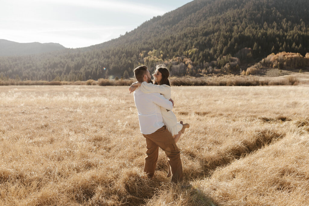 Man hugging and lifting up girlfriend while they look at each other in golden grass field in Lake Tahoe with trees in background