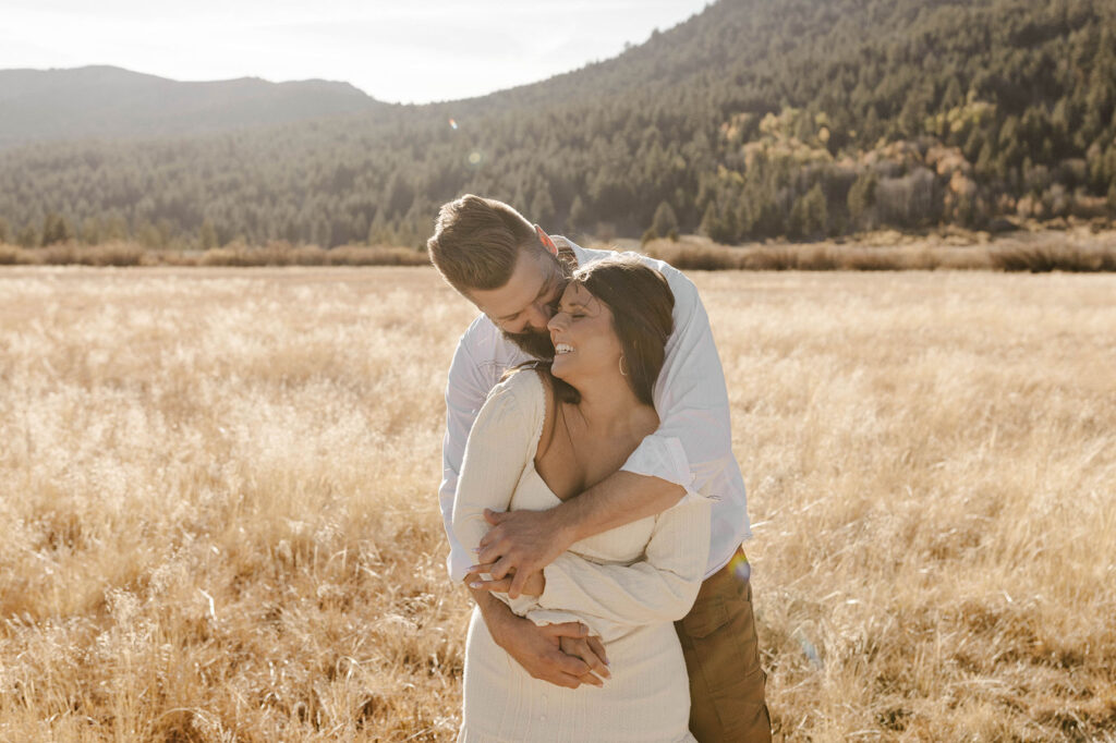 Man leaning over and hugging girlfriend from behind while she smiles and looks back over her shoulder in golden field during Fall in Lake Tahoe